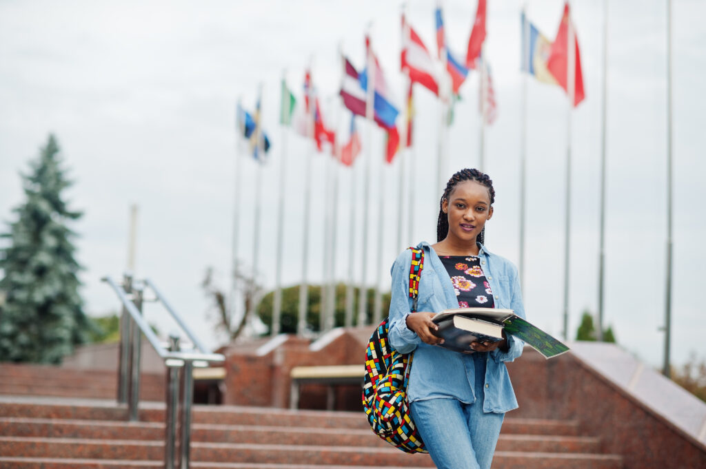 African student female posed with backpack and school items on yard of university, against flags of different countries.